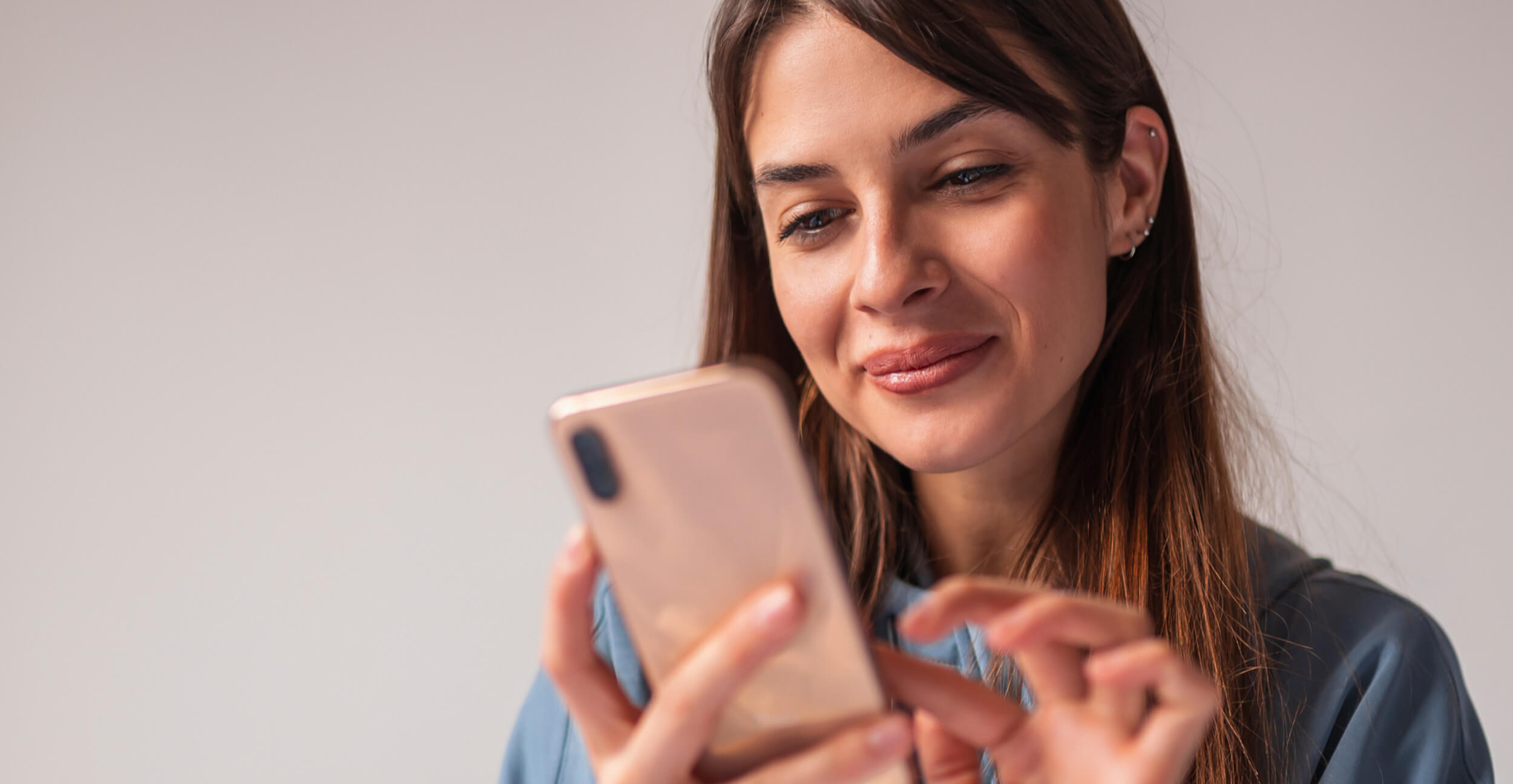 A woman with long brown hair, wearing a blue shirt, holds and uses a smartphone while looking at the screen, smiling slightly.