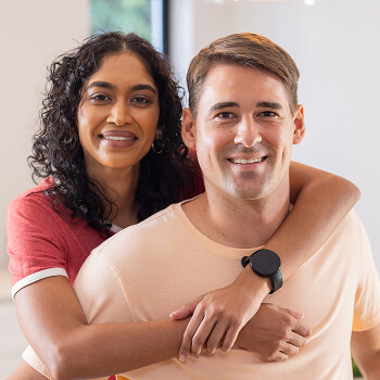 A woman smiles and hugs a man from behind as they pose indoors, both looking at the camera.