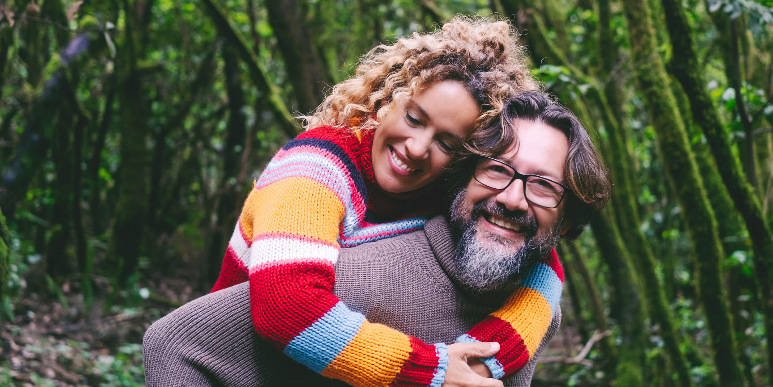A woman in a colorful sweater smiles as she hugs a man from behind in a forest. The man, wearing glasses and a brown sweater, also smiles.