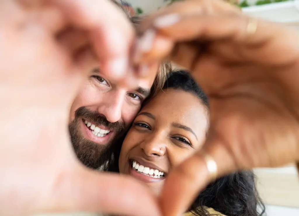 A smiling couple poses closely together, framing their faces with their hands in the shape of a heart.