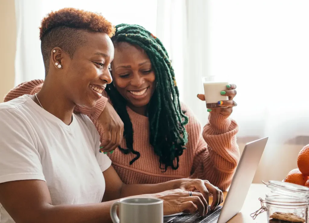 Two women sit together at a table, smiling while looking at a laptop. One is holding a glass of milk and resting her arm on the other's shoulder. A jar and fruit are on the table.