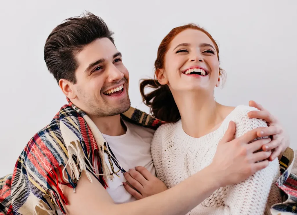 A smiling man and woman sit close together, wrapped in a plaid blanket against a plain white background.