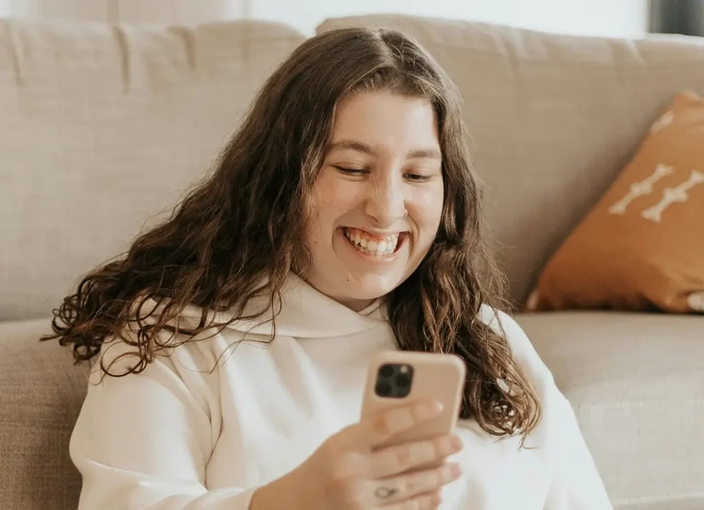 A woman with long brown hair smiles while looking at her smartphone, sitting in front of a beige couch.